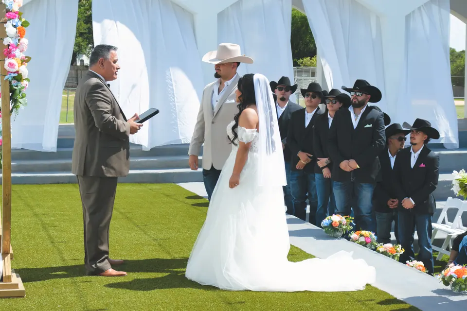 Novios intercambiando votos bajo un arco floral dorado con los padrinos de sombrero vaquero en el Phoenician Ballroom Event Center en San Antonio