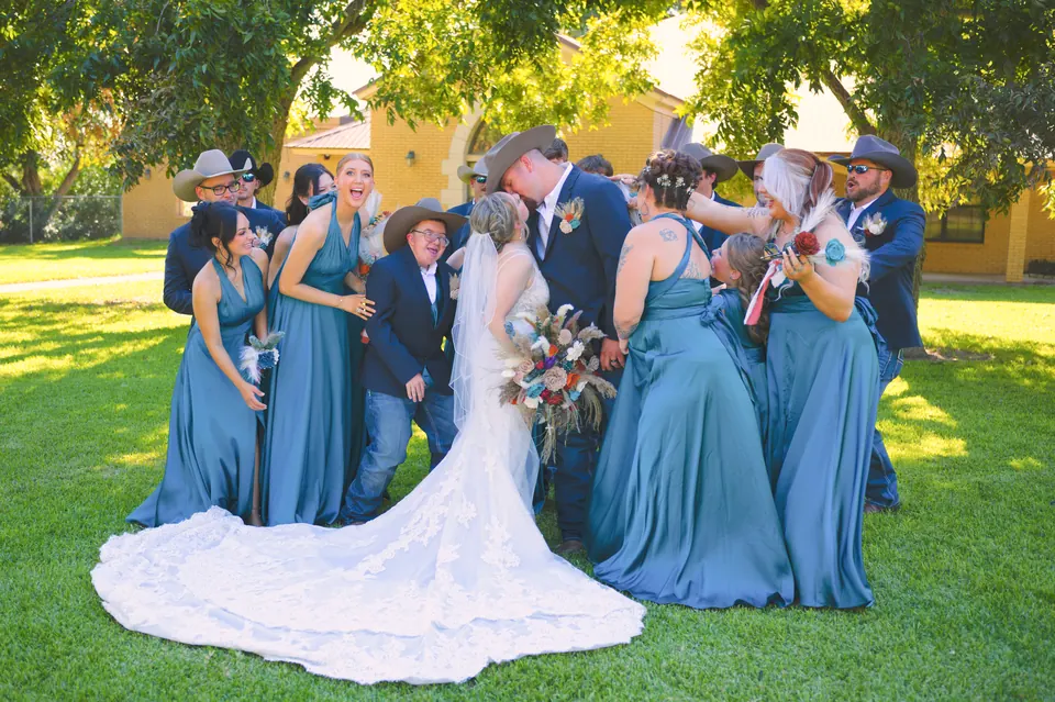 Bridesmaids in dusty blue gowns and groomsmen in cowboy hats laughing around the couple in Kosciusko, Texas