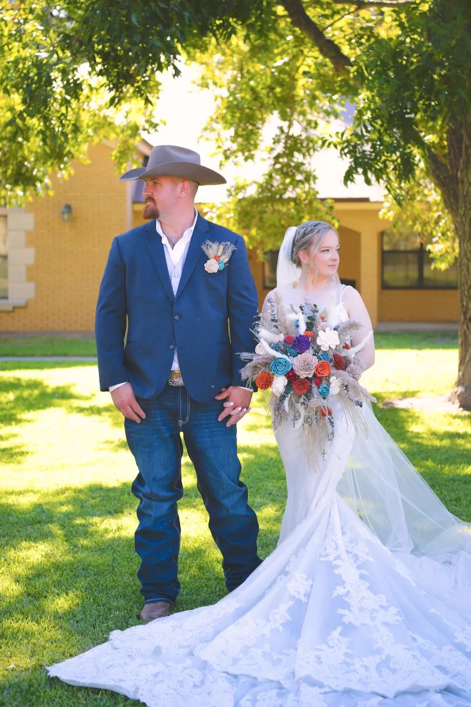 Groom in a cowboy hat and navy blazer beside the bride holding a dried-flower bouquet under oak trees in Kosciusko, Texas