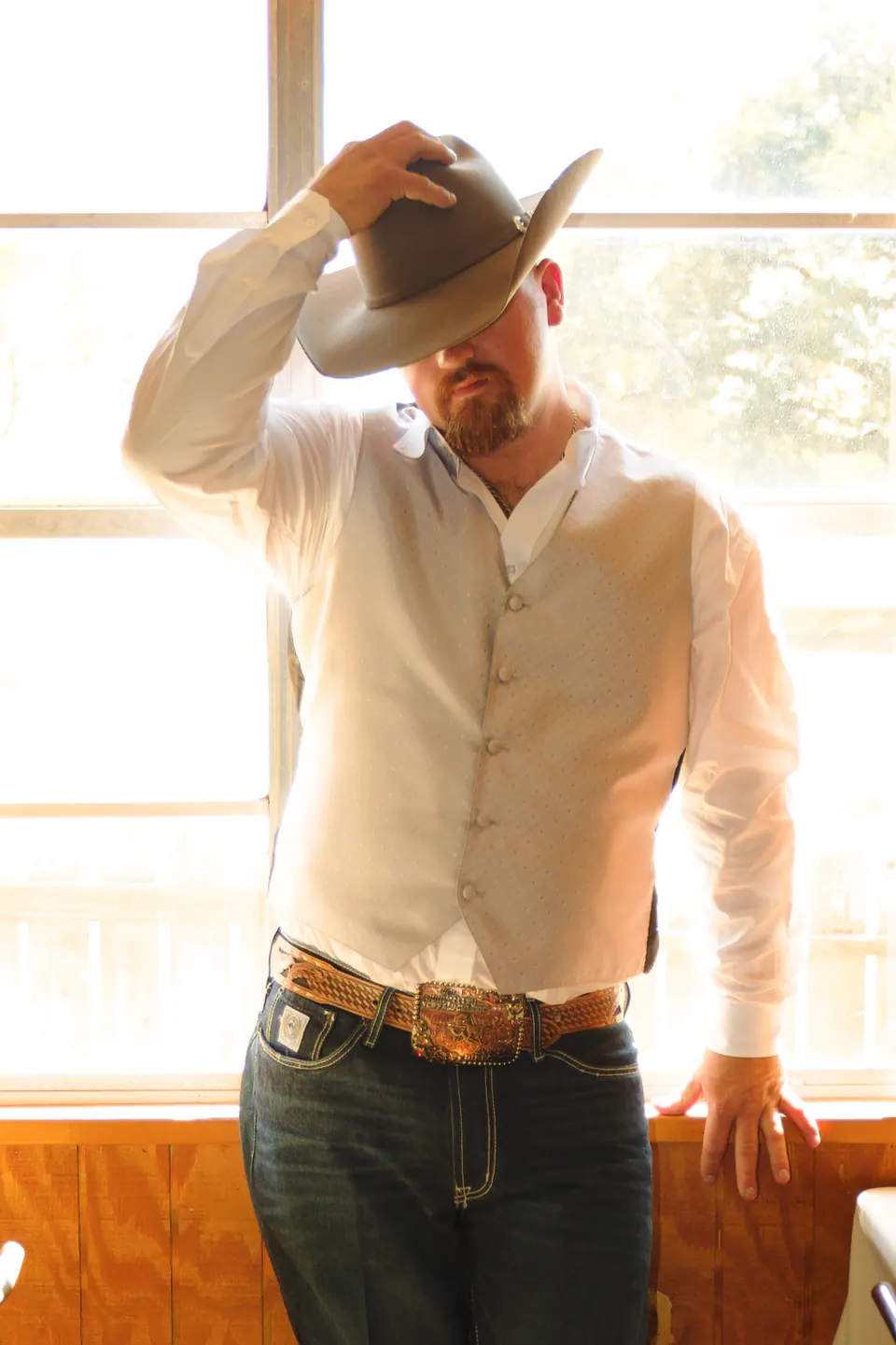 Groom tipping his cowboy hat in a sunlit window before his Kosciusko, Texas wedding