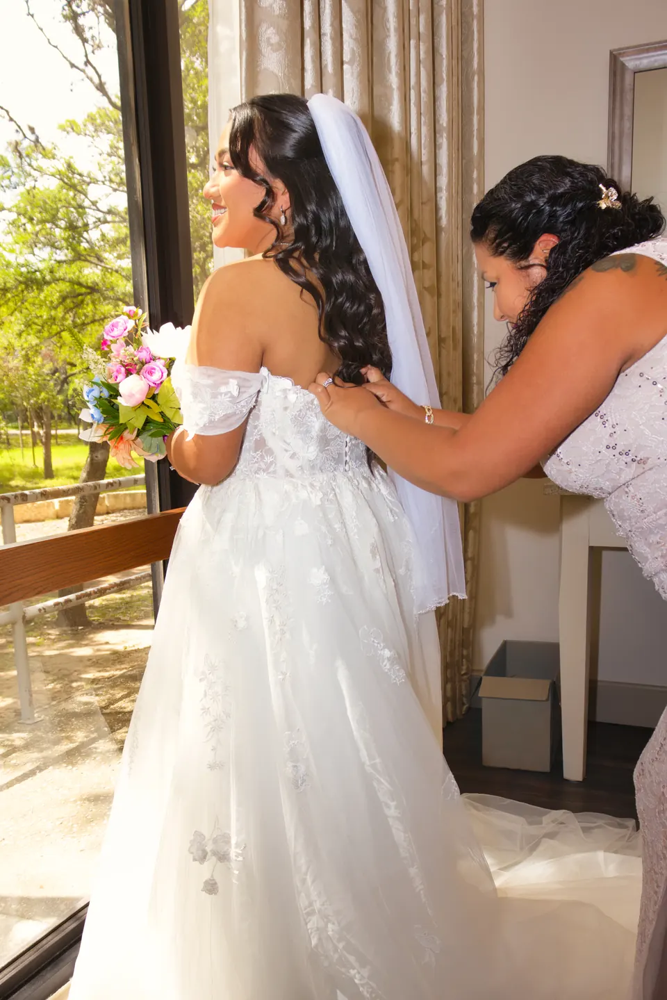 Bride getting help with her gown by the window while getting ready for her San Antonio wedding