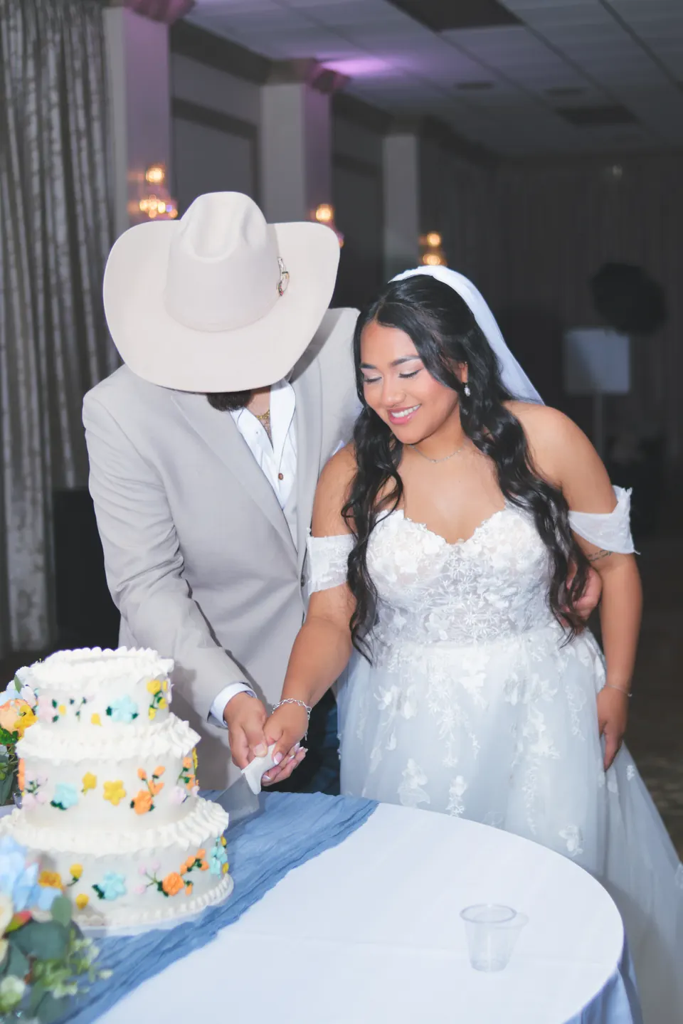 Bride smiling while cutting a floral wedding cake with the groom at the Phoenician Ballroom Event Center in San Antonio