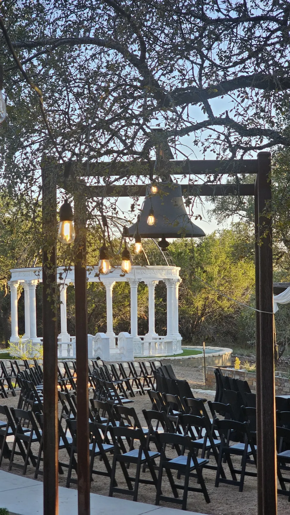 Bell hanging from a timber frame with string lights, framing the marble dome above the ceremony seating at Prima Vista in Wimberley