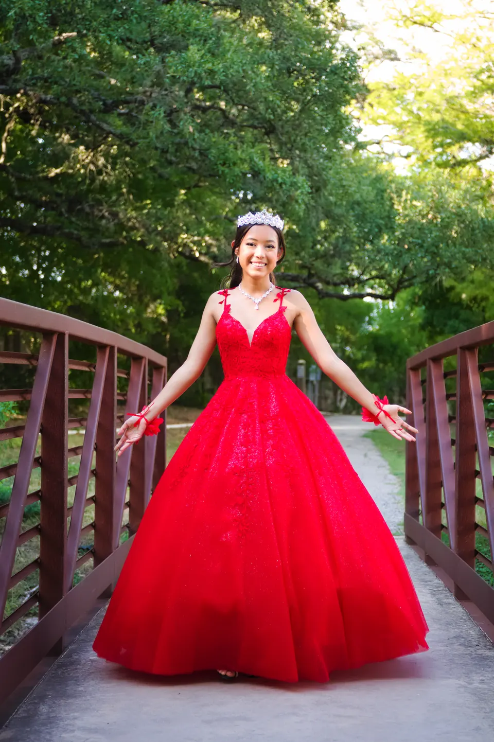 Quinceañera with arms open on a wooden footbridge framed by green trees at Denman Estate Park in San Antonio