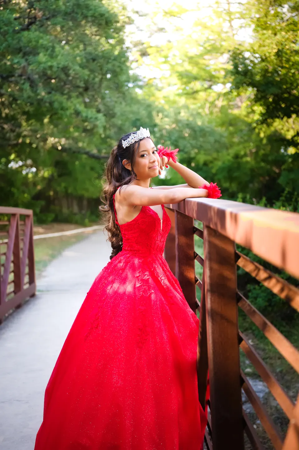 Quinceañera recargada en el barandal de un puente entre árboles verdes en Denman Estate Park, San Antonio