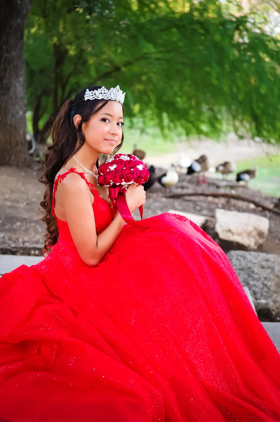 Quinceañera seated by a pond with her bouquet and red ball gown at Denman Estate Park in San Antonio