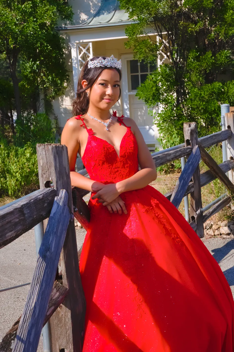 Quinceañera junto a una cerca de madera con su vestido rojo y tiara en Denman Estate Park, San Antonio