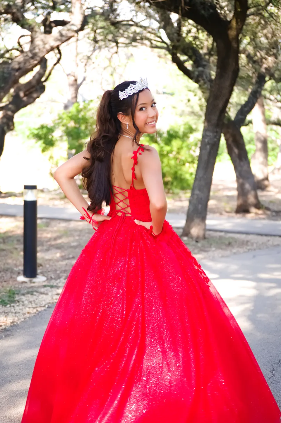 Quinceañera looking over her shoulder to show the corset back of her red gown under oak trees at Denman Estate Park in San Antonio