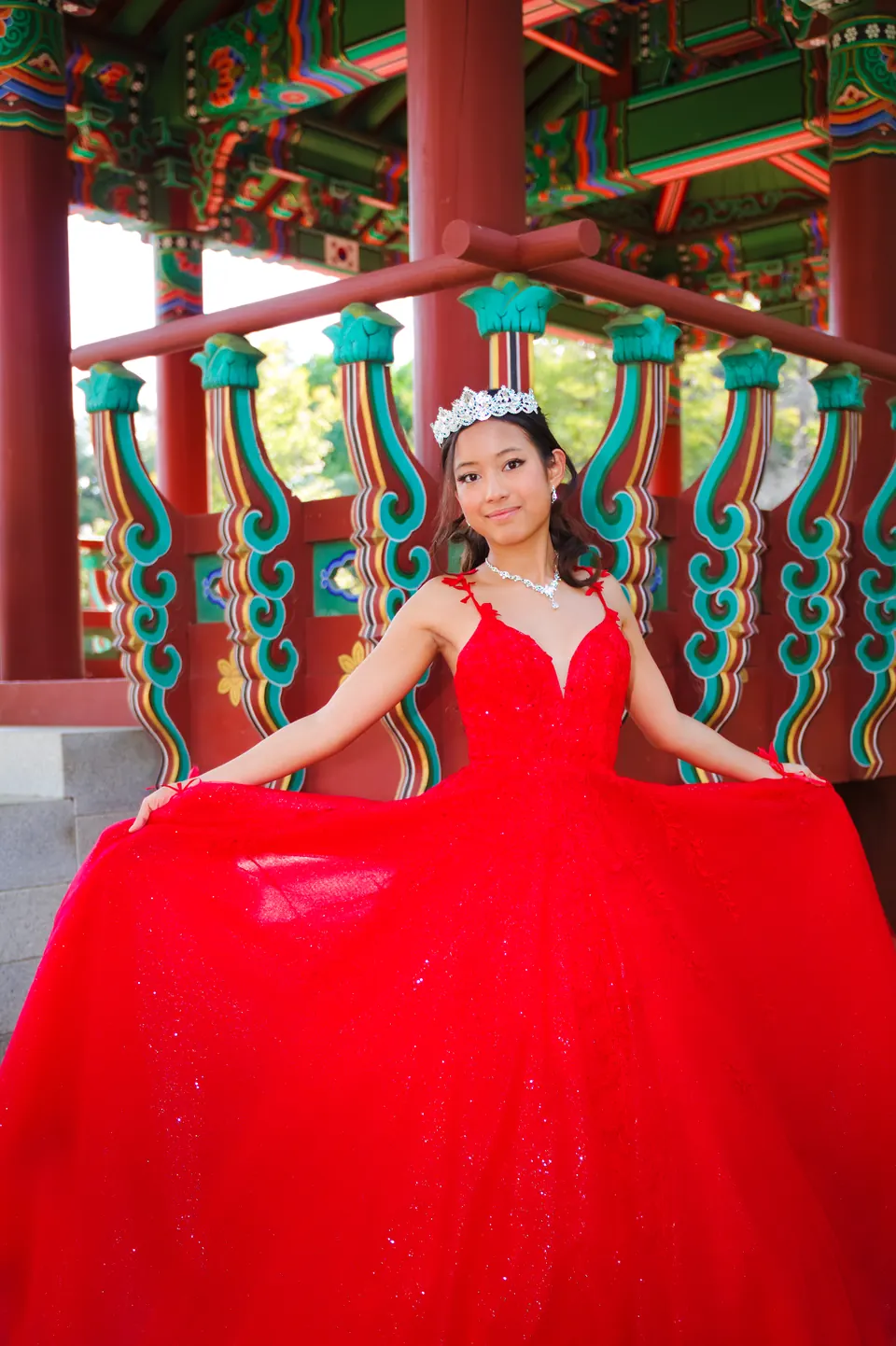 Quinceañera extendiendo la falda de su vestido rojo frente al pabellón coreano en Denman Estate Park, San Antonio