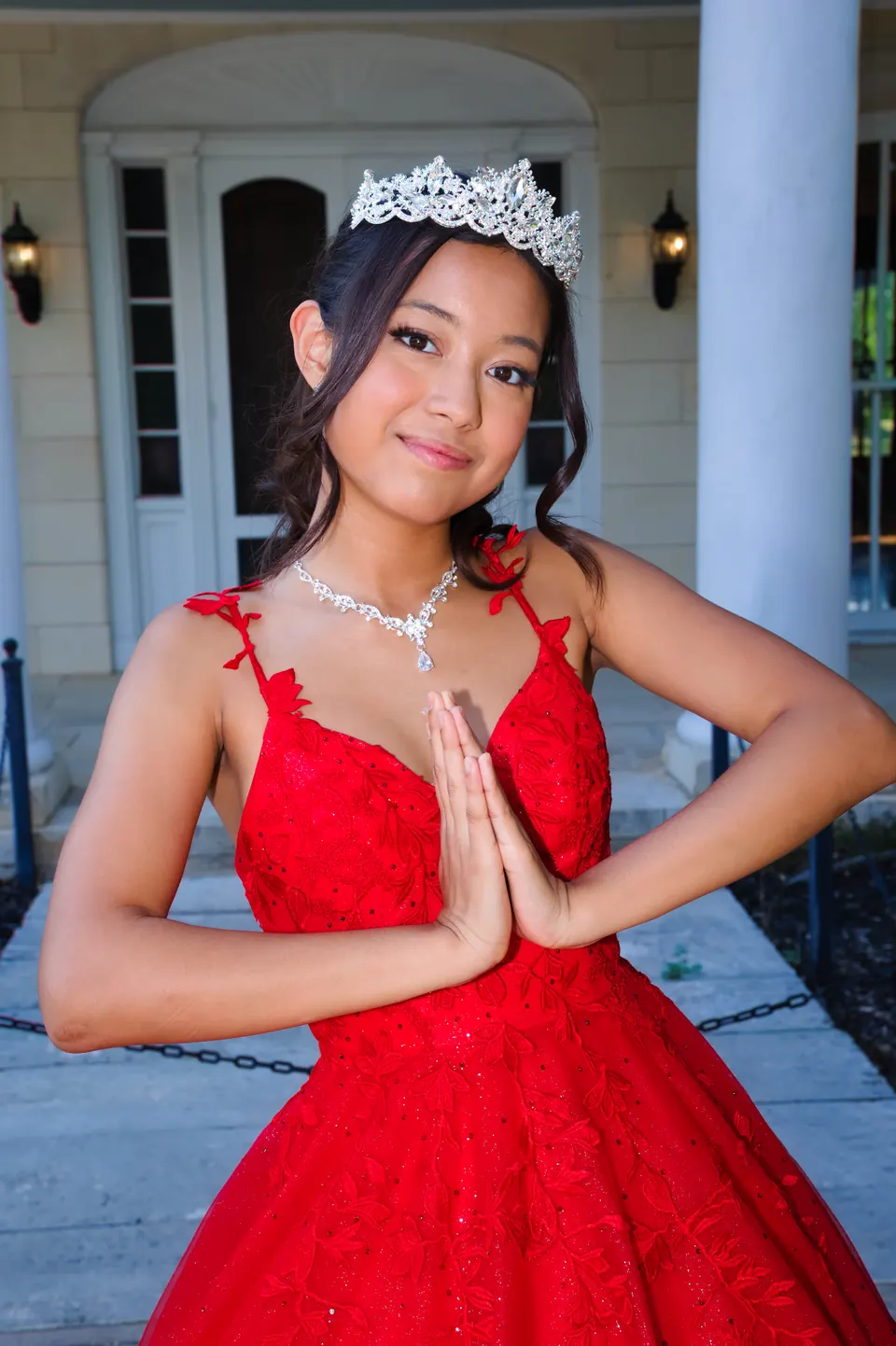 Close-up of a smiling quinceañera in a tiara and red gown with her hands pressed together at Denman Estate Park in San Antonio