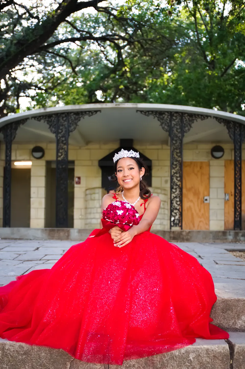 Quinceañera sentada con su ramo de rosas frente a un pabellón de piedra en Denman Estate Park, San Antonio