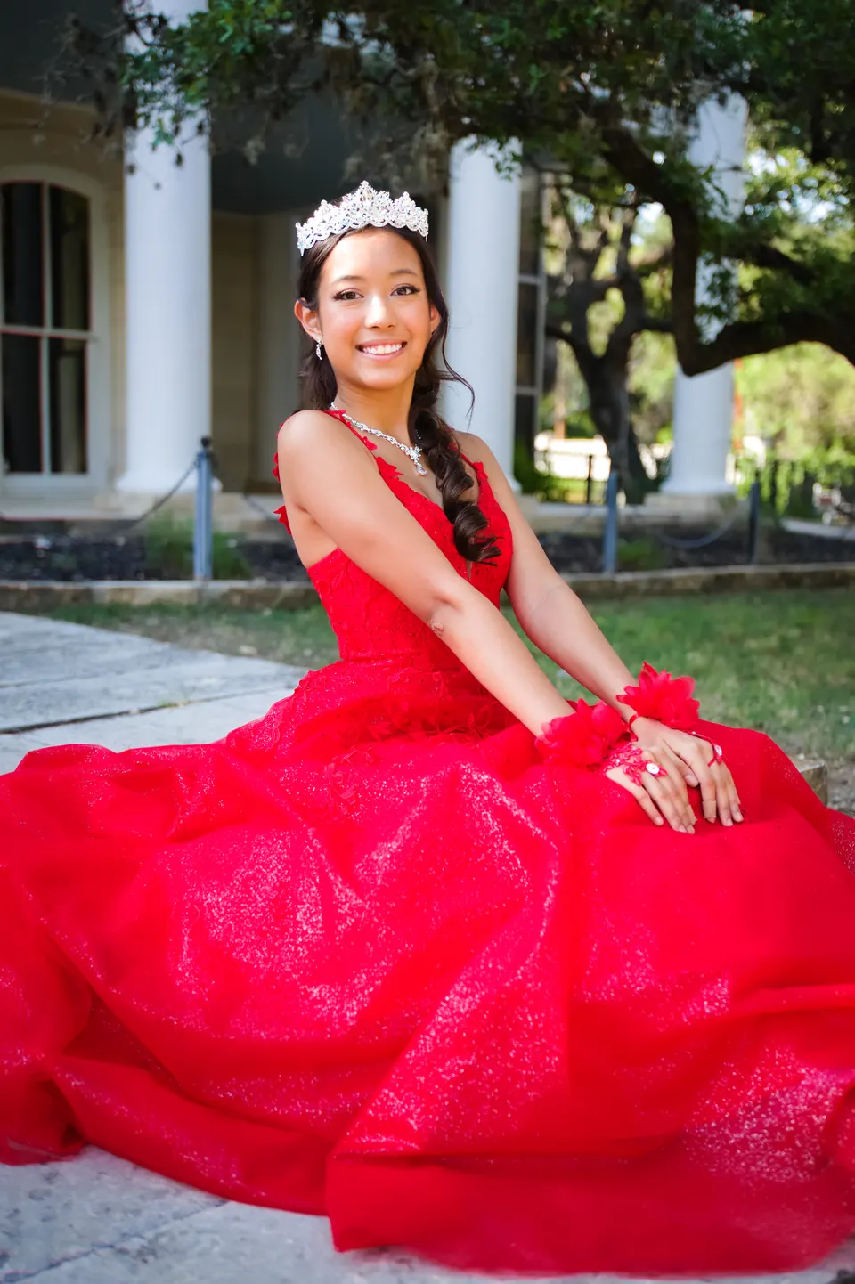 Quinceañera seated with her red gown spread across the lawn beside a mansion at Denman Estate Park in San Antonio