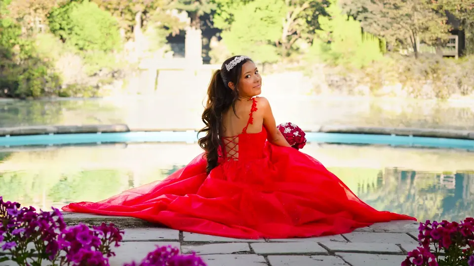 Quinceañera in a red ball gown and tiara seated by a fountain during her pre-quince portraits at Denman Estate Park in San Antonio