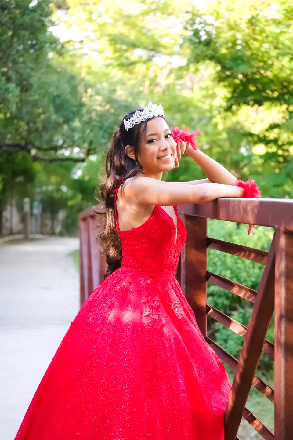 Quinceañera leaning on a wooden railing in her red gown and tiara for a pre-quince portrait at Denman Estate Park in San Antonio